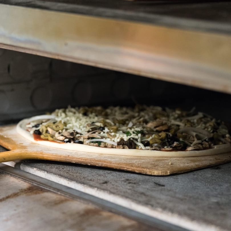 Uncooked Genova's pizza with mushrooms, olives, and cheese on a wooden peel entering a hot oven.
