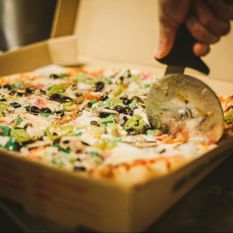 A hand uses a pizza cutter to slice a Genova's Italian Restaurant pizza with various toppings.