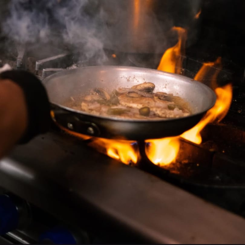A chef's hand holds a pan of chicken and mushrooms cooking over a fiery stove.