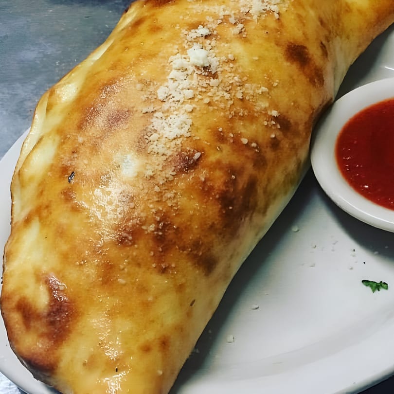 Golden-brown calzone sprinkled with parmesan cheese, served with a side of red marinara sauce.